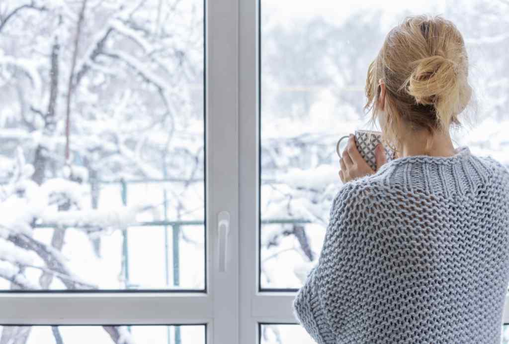 A woman looks out of the window at the snow-covered outdoors during a furnace installation with a cup of drink in her hands.